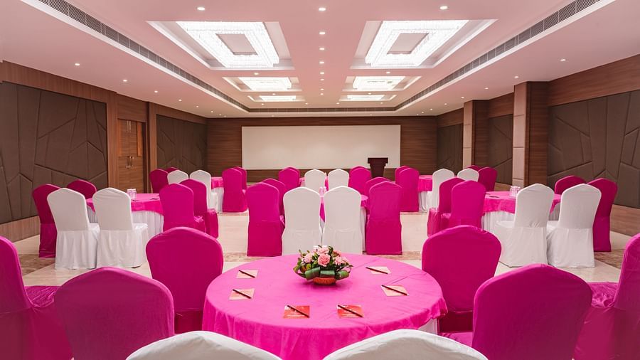 A banquet hall at Lords Inn Udaipur is set for an event with round tables draped in pink, surrounded by chairs in pink and white covers, facing a stage.