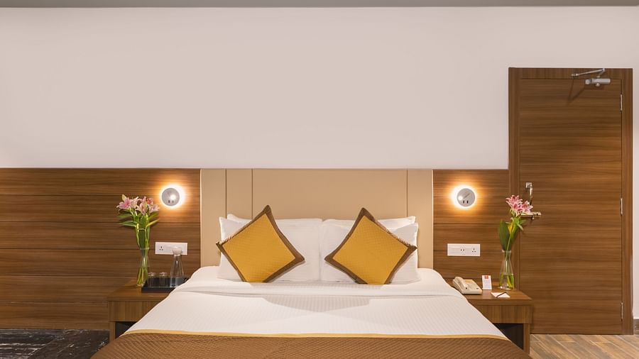 A comfortable hotel room at Lords Inn Udaipur features a double bed with brown accents, flanked by bedside tables with lamps and flowers.