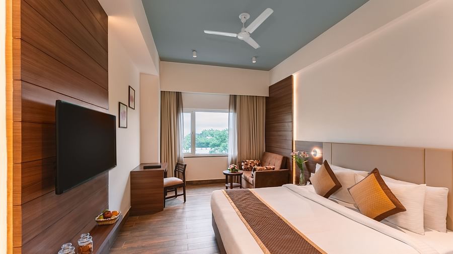 A well-lit hotel room at Lords Inn Udaipur offers a comfortable bed, a seating area by the window, a wall-mounted TV, and a ceiling fan.
