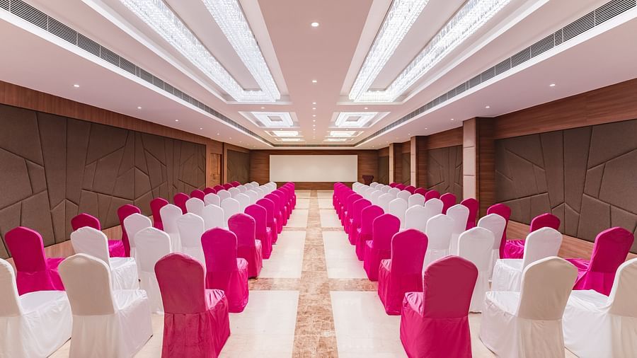 A large banquet hall at Lords Inn Udaipur is arranged with rows of tables and chairs draped in white and vibrant pink fabric, facing a stage.