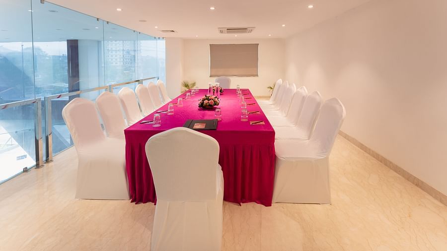 A modern meeting room at Lords Inn Udaipur is arranged with a long table draped in a bright pink cloth, surrounded by white chairs, and featuring water glasses and floral accents.