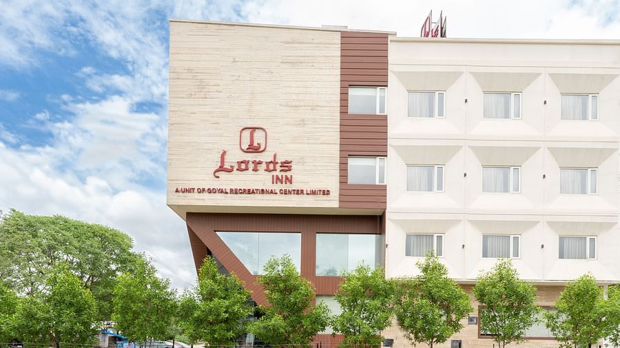 The Lords Inn Udaipur hotel stands under a bright, partly cloudy sky, surrounded by green trees.