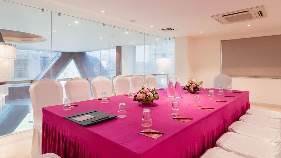 A meeting room at Lords Inn Udaipur is set with a long table covered in a vibrant cloth, surrounded by white chairs and adorned with floral centerpieces and water glasses.