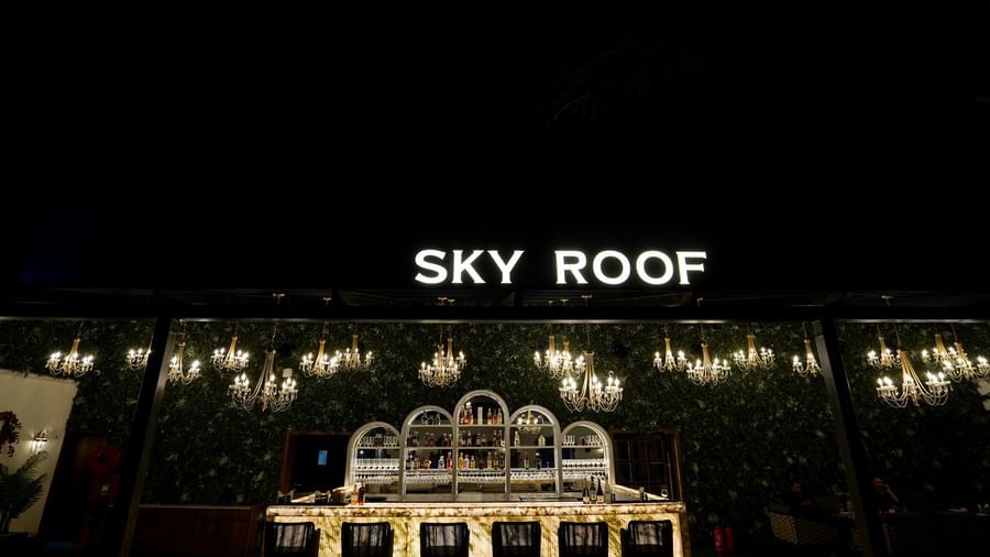 A wide angle shot of the Sky Roof featuring a brightly lit bar area along with a neon sign board at the top at M2 Square by Monday Premium.