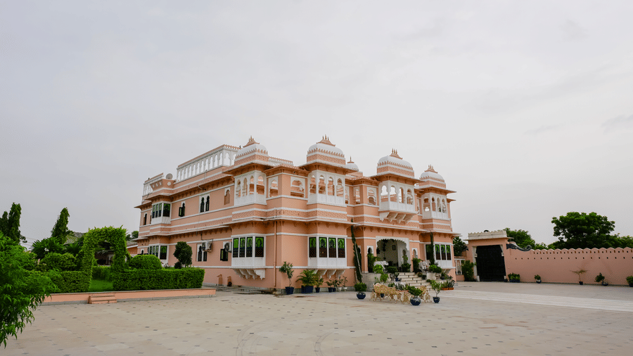 A panoramic ground-level shot of the front of Mahendra Niwas, highlighting the stunning pale pink facade, with a lush lawn and a clear sky in the background.