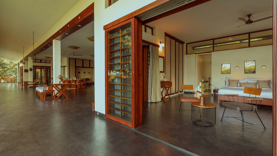 Villa Wuwei at Mango Hill Auroville, Pondicherry featuring a bedroom with a bed and chairs opens onto a wide veranda with pillars and a dining table through large sliding doors.