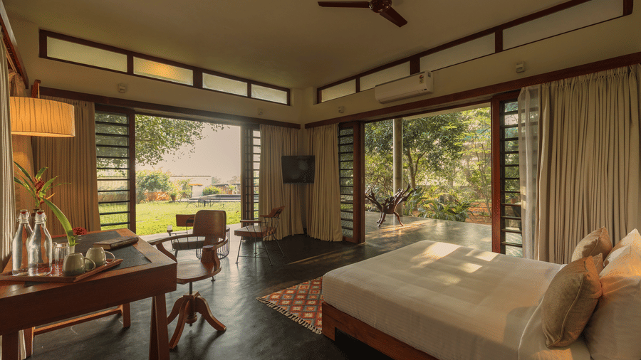 Villa Wuwei in Mango Hill Auroville, Pondicherry featuring a bed is placed near corner windows with curtains and a desk with a chair is positioned nearby.
