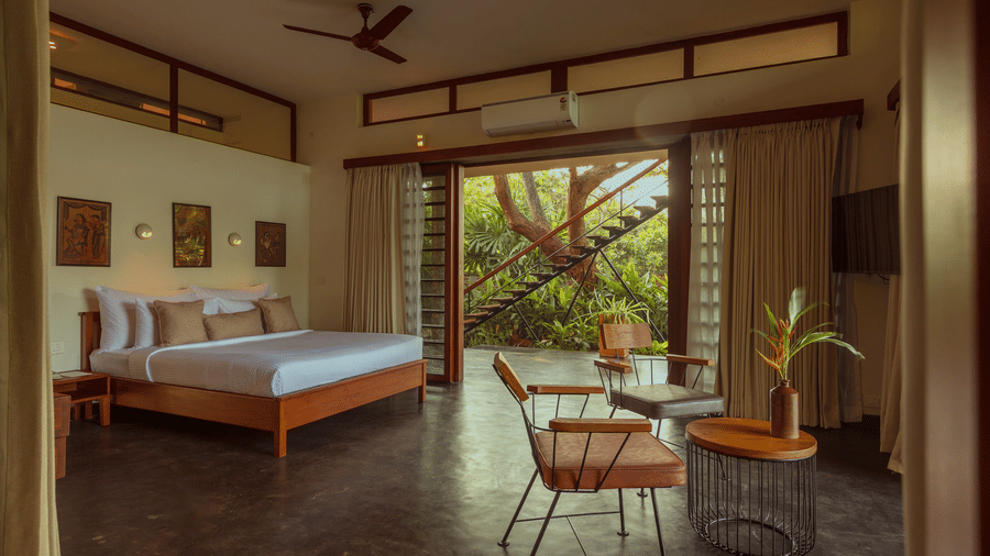 A bed at Villa Wuwei in Mango Hill Auroville, Pondicherry faces an open doorway leading to a veranda with chairs and a table, with trees visible outside.