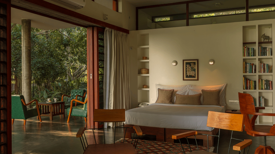 Villa Wuwei at Mango Hill Auroville, Pondicherry featuring a bedroom features a double bed, a built-in bookshelf, and wooden chairs facing an open sliding door.