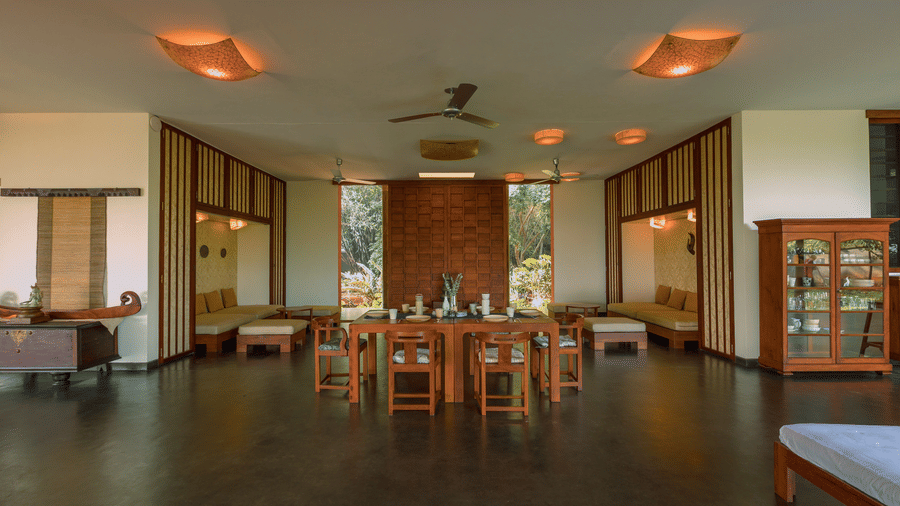 A dining area Villa Wuwei in Mango Hill Auroville, Pondicherry featuring a long table and chairs is set in a hall with ceiling fans and windows on both sides.