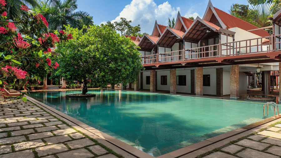 A sunlit view of the turquoise swimming pool and traditional Thai-style villas at Mango Hill Auroville, Pondicherry.