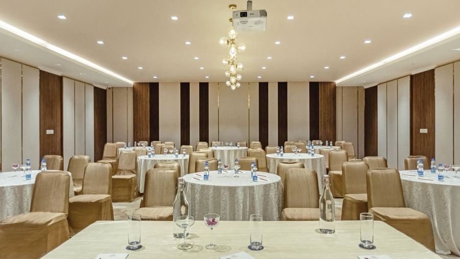Banquet hall at Mango Hill Central, Chennai, with round tables and chairs arranged for a meeting or event. A projector hangs from the ceiling.