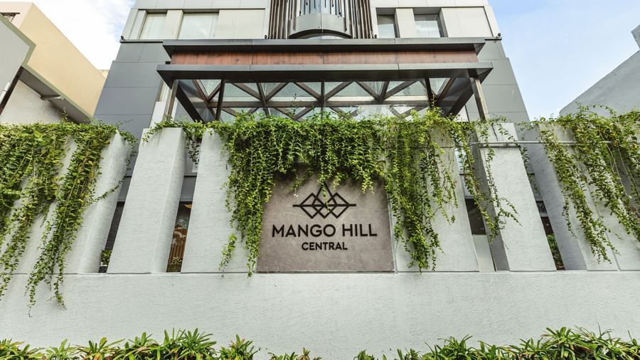 An exterior view of Mango Hill Central, Chennai, with lush green plants on the facade and the hotel name visible.