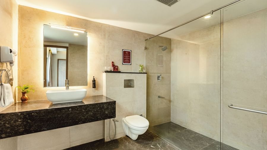 A wide-angle view of the bathroom at Mango Hill Central, Chennai, showing the sink, mirror, toilet, and glass-enclosed shower.