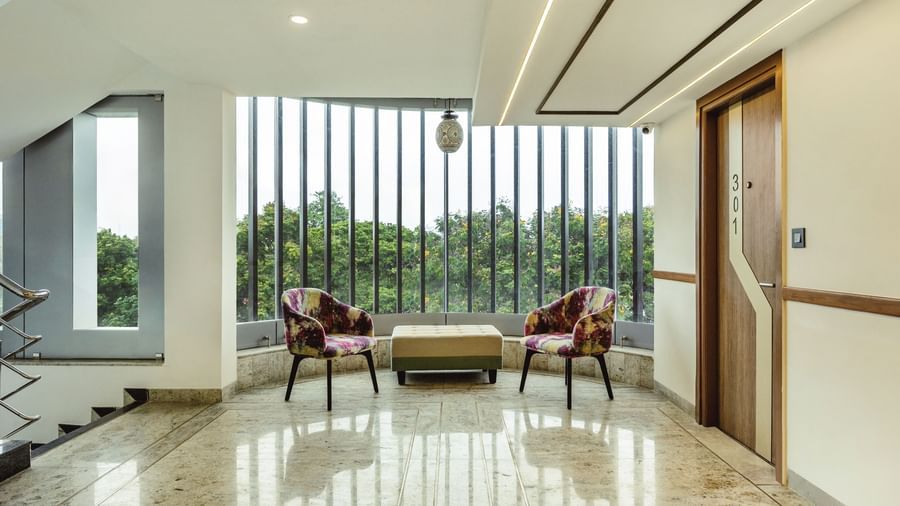 A seating area with a sofa and two chairs at Mango Hill Central, Chennai. Large windows with a view of greenery are visible.