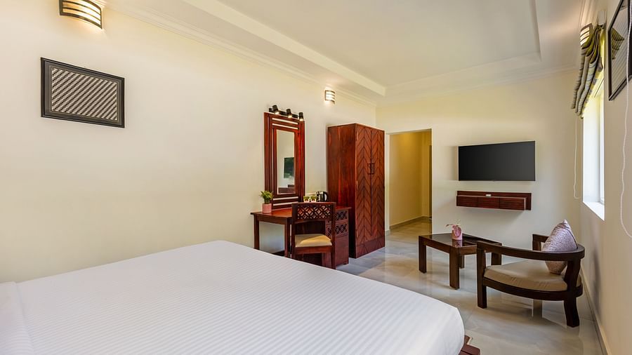 A well-lit hotel room with a bed, a desk, a wardrobe, a television, and a seating area - Mango Hill Resort, Coimbatore