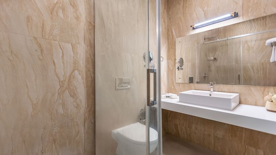 A bathroom with beige and brown textured walls, a glass shower stall, a white rectangular sink, and a mirror - Mango Hill Resort, Coimbatore