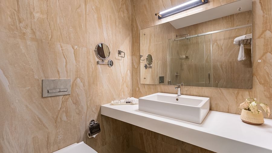 A bathroom with beige and brown textured walls, a white rectangular sink, a large mirror, and a white toilet - Mango Hill Resort, Coimbatore