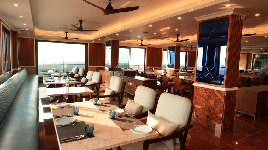 Indoor restaurant seating with cushioned chairs, tables set for dining, and ceiling fans overhead at MAYFAIR Bay Resort, Paradeep.