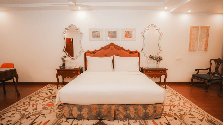 Deluxe suite with a king bed, carved tufted headboard, bedside tables with a telephone and flowers, framed artwork and mirrors at MAYFAIR Garden, Rourkela.