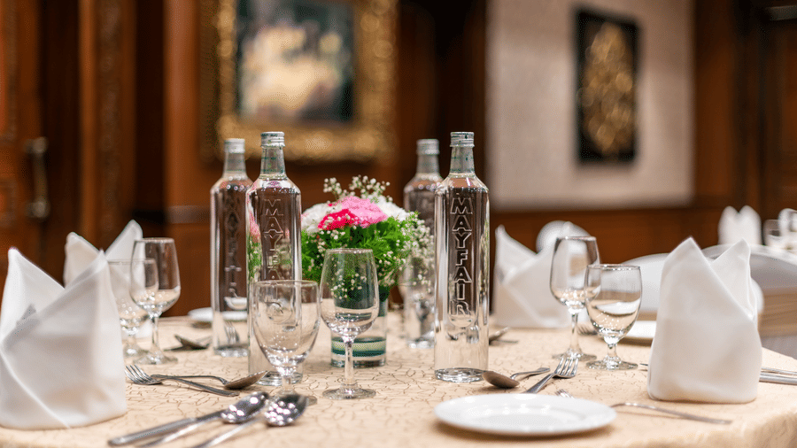 Round table laid with white napkins, glassware, embossed water bottles, cutlery and a floral centrepiece in Orchid banquet hall at MAYFAIR Garden, Rourkela.
