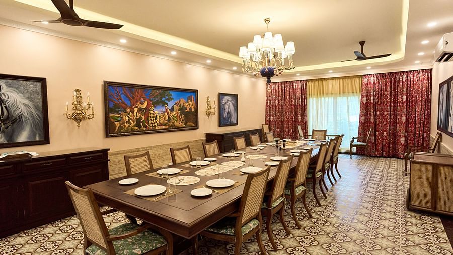 Diagonal view of a large dining table set for a culinary experience, with wall-mounted paintings, a chandelier, and large windows at Mayfair Manor, Alibag