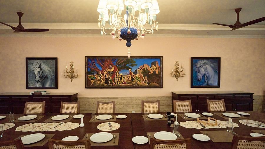 A large dining table elegantly set for a culinary experience, complemented by wall-mounted paintings and a luxurious chandelier overhead at Mayfair Manor, Alibag