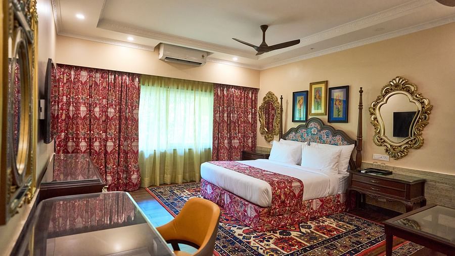 A spacious room featuring a large bed, a desk with a chair, wooden bedside tables, and windows draped with curtains at Mayfair Manor, Alibag