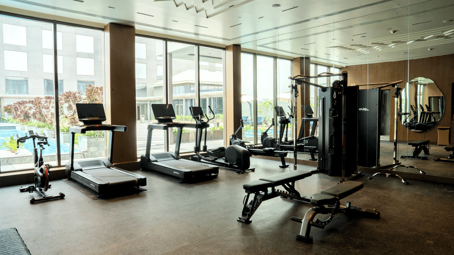 Modern gym with treadmills, exercise bike, elliptical, weight bench, and multi-gym machine, large windows overlooking pool and plants, and mirrored wall reflecting equipment and natural light.