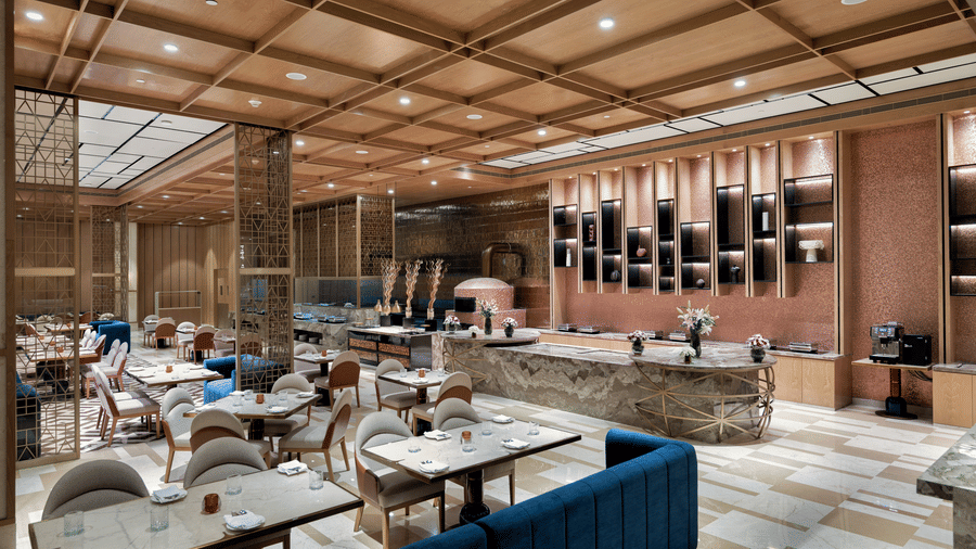 Interior view of Mercano restaurant at Merlis Hotel, Coimbatore showing dining tables with chairs, sofas, place settings, a service counter, wall shelves, lighting fixtures, and a ceiling with wooden panels.