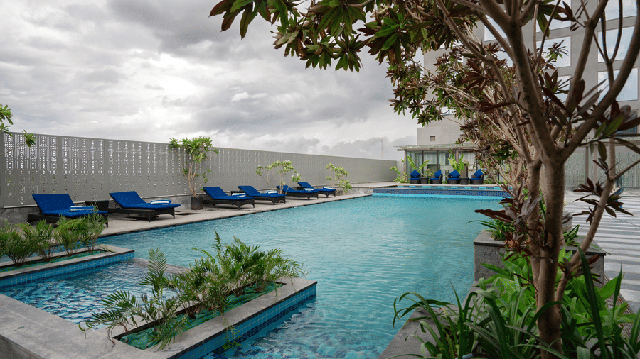 Outdoor swimming pool at Merlis Hotel, Coimbatore, featuring blue loungers, landscaped greenery, and a modern poolside setting under a cloudy sky.
