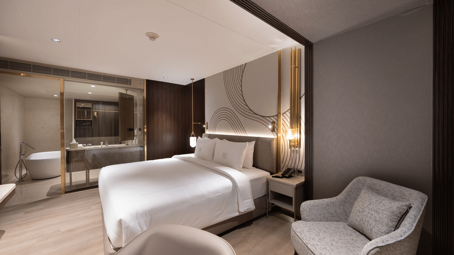 A hotel room with a king size bed, stylish wall art, bedside tables with lamps, a grey armchair, and a glass-partitioned bathroom featuring a freestanding bathtub and vanity - Merlis Hotel, Coimbatore
