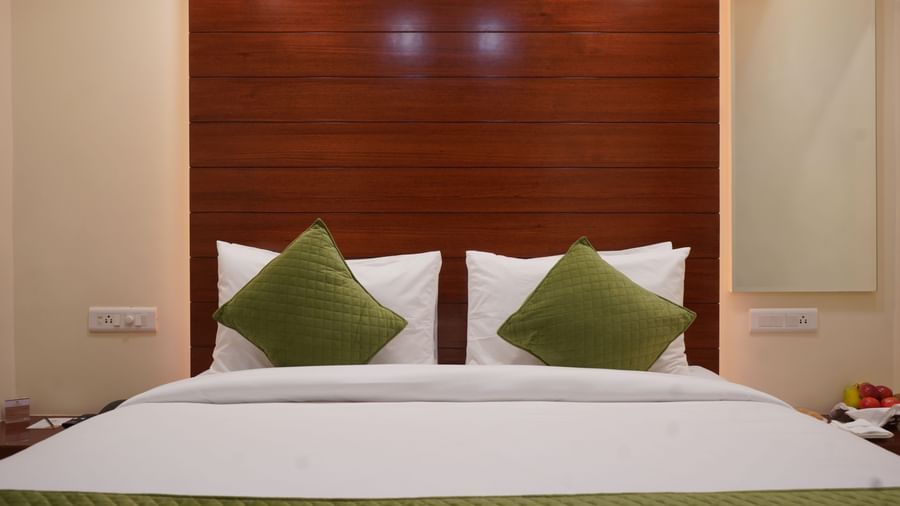 A wide shot of a hotel room with two single beds, each with a white comforter and a green accent runner. The beds are separated by a small table.
