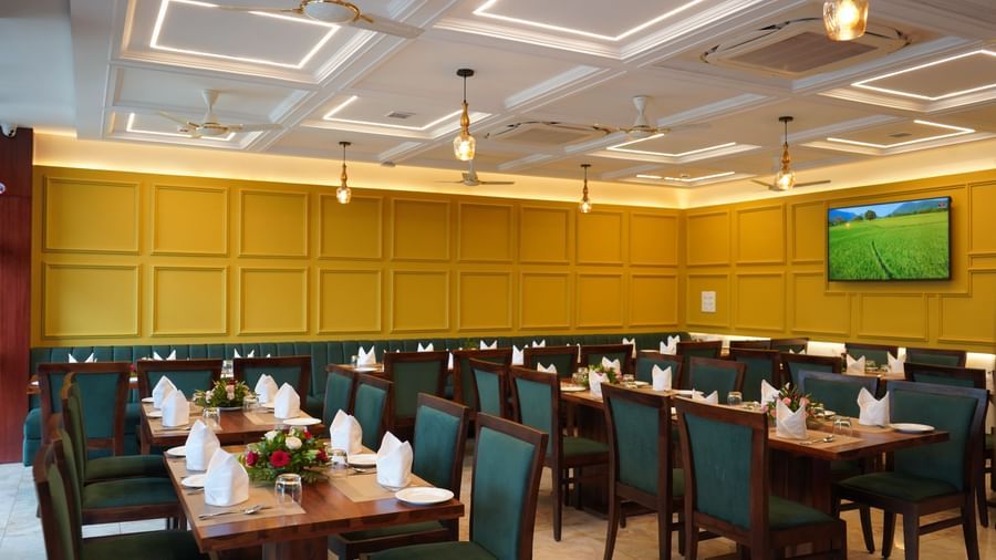 A wide shot of a dining hall with multiple tables set for a meal. The chairs are dark green, and the walls are yellow. A TV is visible on the far wall.