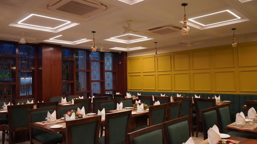 A wide shot of a dining hall with multiple tables set for a meal. The walls are yellow, and the chairs are dark green. The ceiling has multiple recessed lights.
