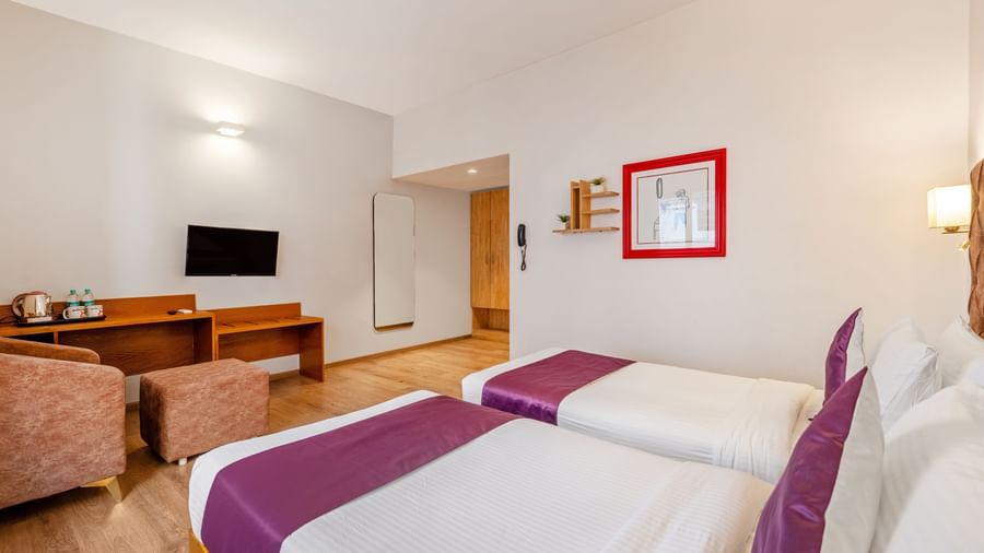 A spacious hotel room with 2 single beds, a TV on top of a wooden desk, a chair, a mirror, and complimentary water bottles – Monday Hotels HITEC City, Hyderabad