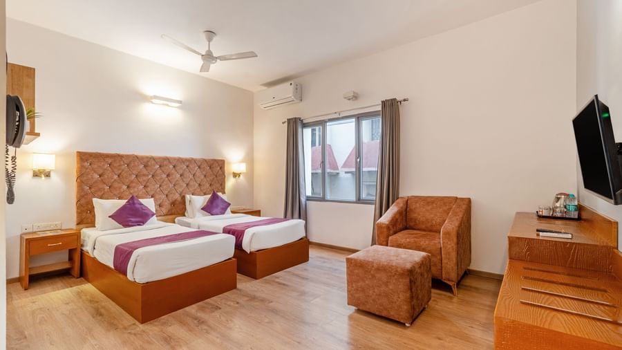 A bright hotel room with 2 single beds, cushioned headboards, a TV, a wooden desk, chairs, and a large window – Monday Hotels HITEC City, Hyderabad
