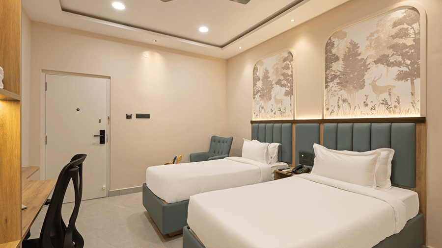 Twin beds with green padded headboards below forest murals, armchair, work desk and entrance door in a Deluxe room at Monday Hotels & Suites Madhapur, Hyderabad.