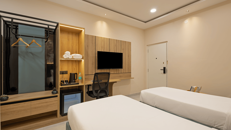 Open wardrobe, towel shelves, kettle, minibar, wall-mounted TV and work desk beside twin beds in a Deluxe room at Monday Hotels & Suites Madhapur, Hyderabad.
