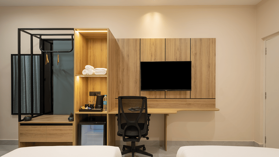 Work desk with chair and wall-mounted TV between twin beds, with an open wardrobe and minibar, in a Deluxe room at Monday Hotels & Suites Madhapur, Hyderabad.