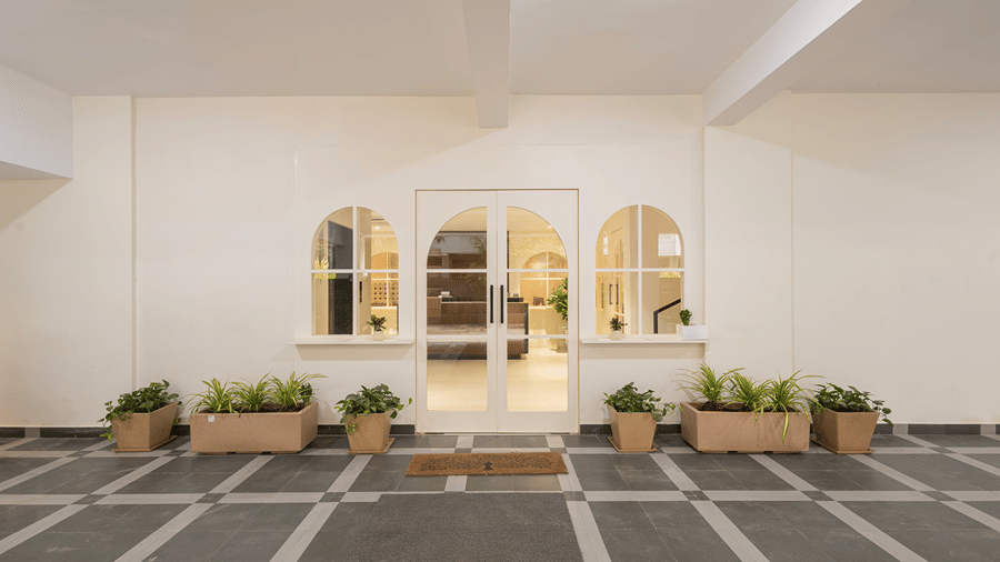 Glass double doors flanked by arched windows and potted plants, with a doormat, at the entrance of Monday Hotels & Suites Madhapur, Hyderabad.