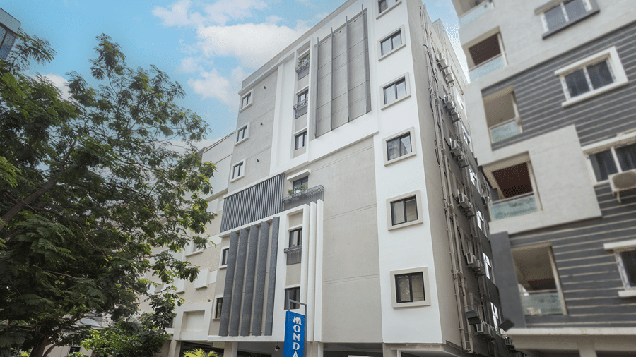 Multi-storey facade with grey and white cladding, a blue vertical sign and a large tree in daylight at Monday Hotels & Suites Madhapur, Hyderabad.
