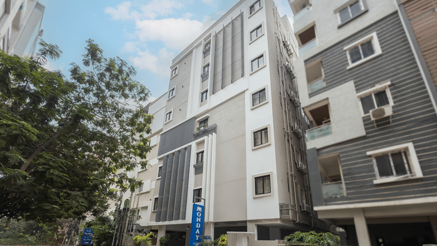 Side angle of the multi-storey facade with a blue vertical sign, trees and neighbouring buildings at Monday Hotels & Suites Madhapur, Hyderabad.