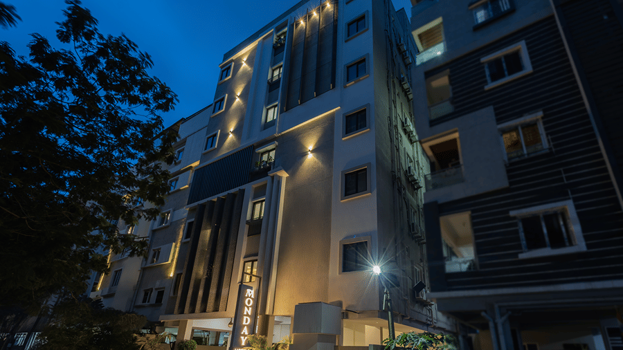 Facade with uplighting on the grey cladding, lit windows, a vertical sign and a street lamp at dusk at Monday Hotels & Suites Madhapur, Hyderabad.