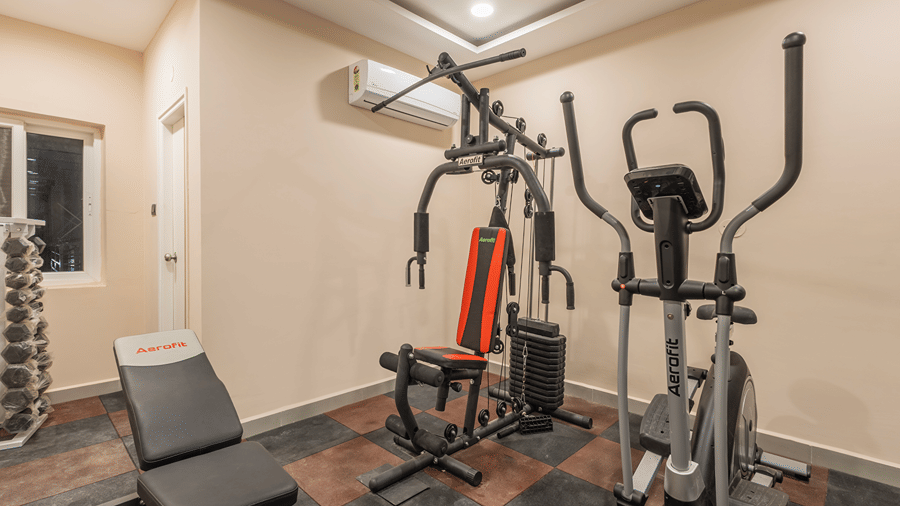 Multi-gym station, elliptical trainer, weight bench and dumbbell rack on chequered flooring in the fitness centre at Monday Hotels & Suites Madhapur, Hyderabad.