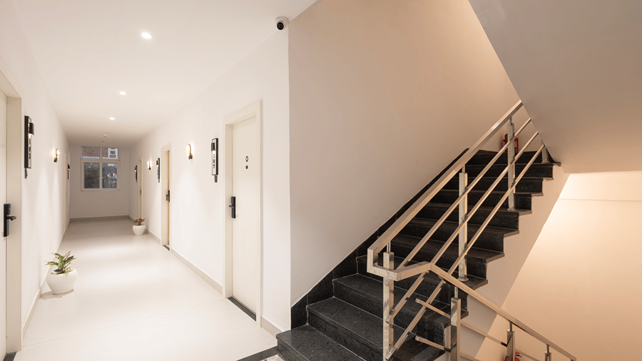 Guest corridor with room doors and potted plants beside a staircase with stainless-steel railings at Monday Hotels & Suites Madhapur, Hyderabad.
