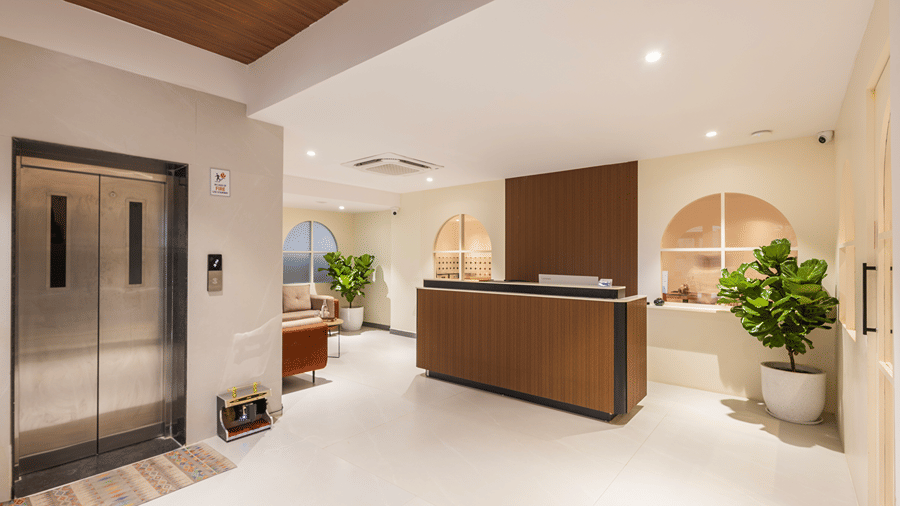 Lobby with lift doors, a shoe-shine machine, patterned rug and the reception desk with potted plants at Monday Hotels & Suites Madhapur, Hyderabad.