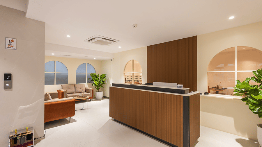 Lobby with a wood-panelled reception desk, sofas, arched windows, potted fiddle-leaf figs and a lift call panel at Monday Hotels & Suites Madhapur, Hyderabad.
