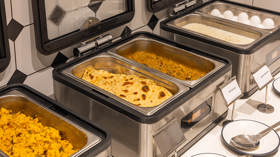 Breakfast buffet chafing dishes with upma, rotis, curry and boiled eggs beside labelled tags at M-Salt at Monday Hotels & Suites Madhapur, Hyderabad.
