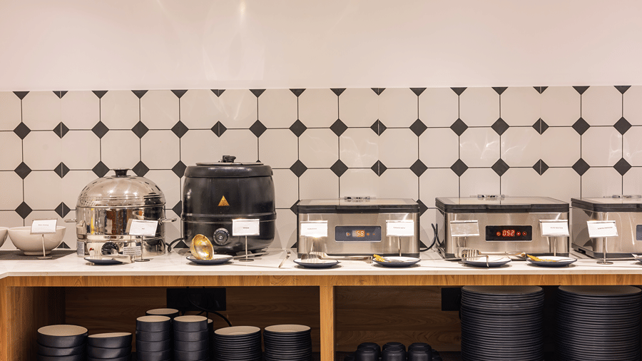 Buffet counter with a steamer, soup kettle and chafing dishes above stacked bowls and plates at M-Salt at Monday Hotels & Suites Madhapur, Hyderabad.